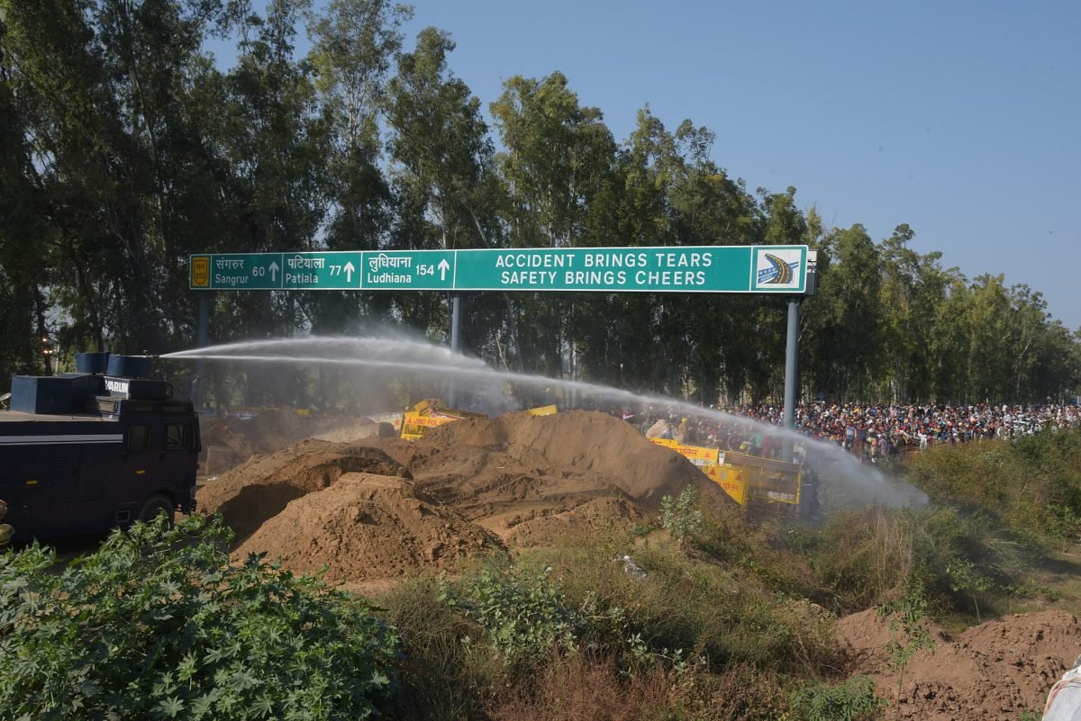 A police water cannon sprays a crowd of farmers beyond heaps of sand piled across a highway, under a sign showing Sangrur 60 km away