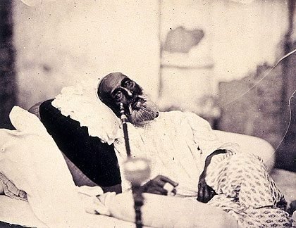An elderly bearded man in a white cap and shawl, lying back on cushions, in an old sepia photograph
