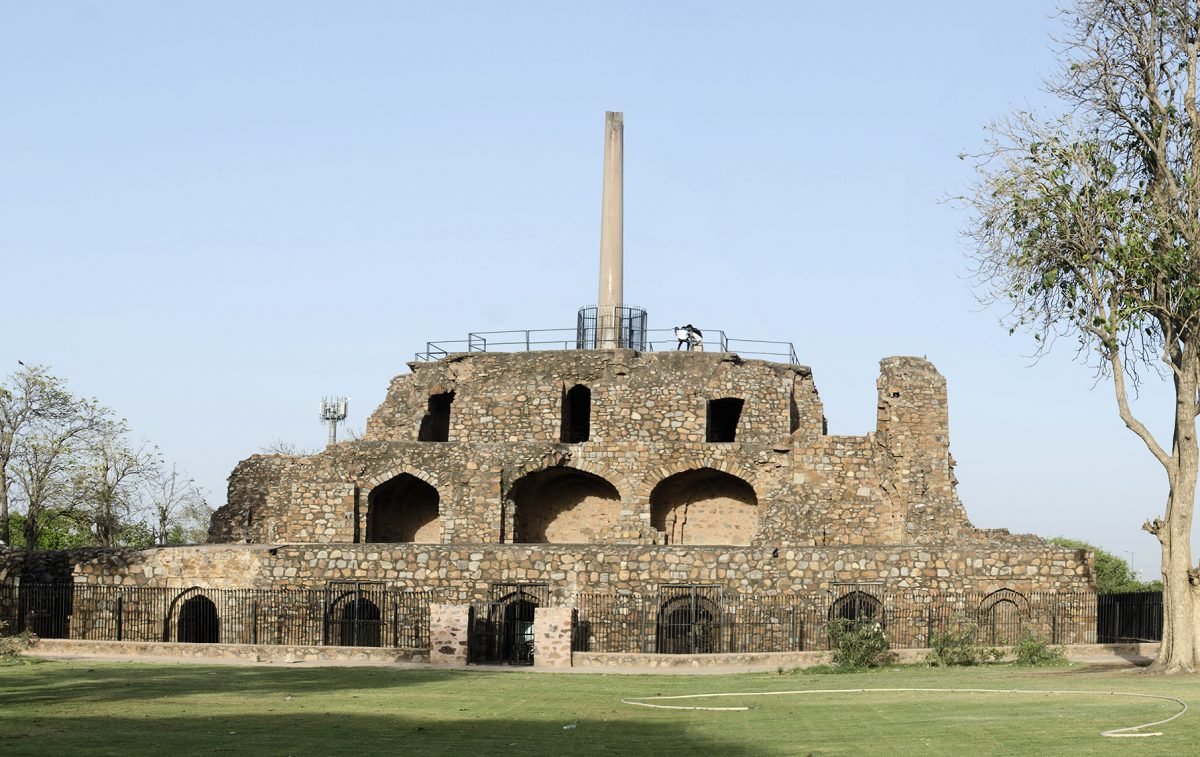 A tall smooth stone pillar standing on top of a ruined stepped stone structure under a clear sky