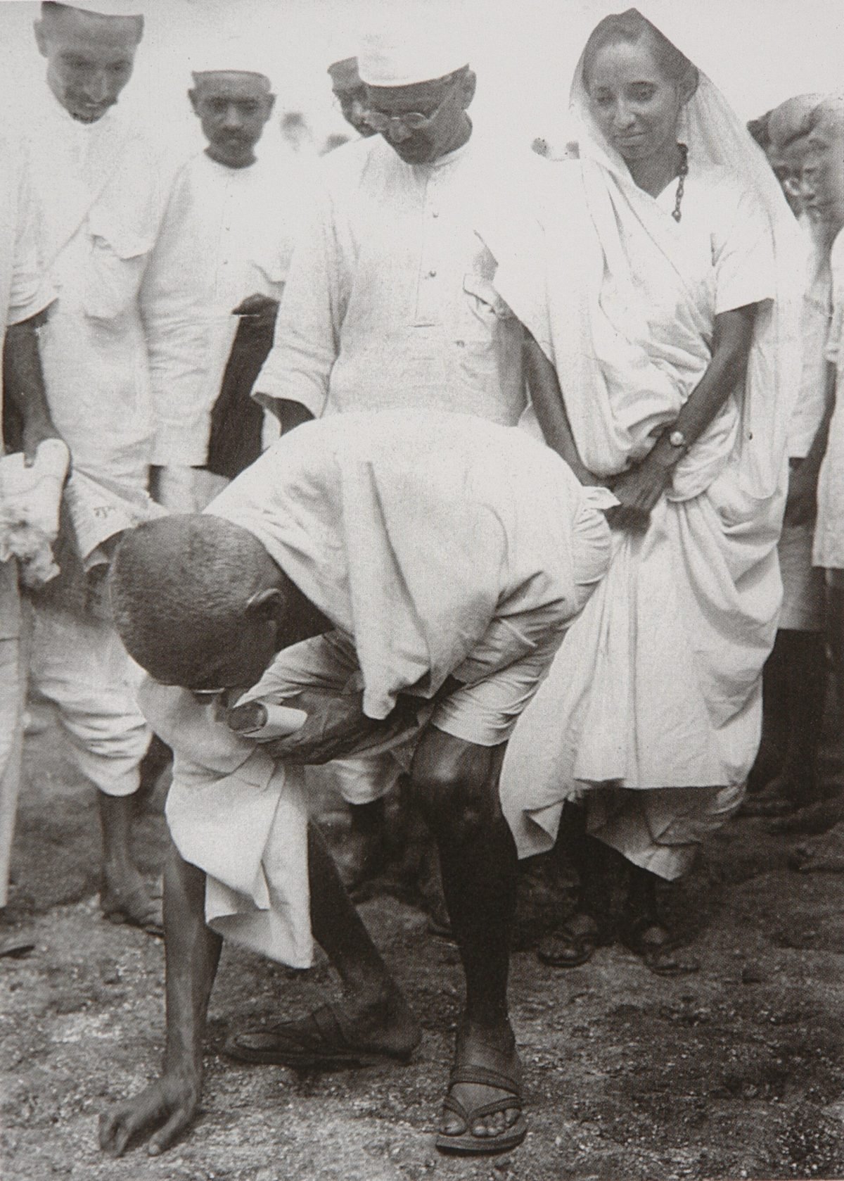 Gandhi bends down on a beach to pick up salt, with a man and a woman standing behind him