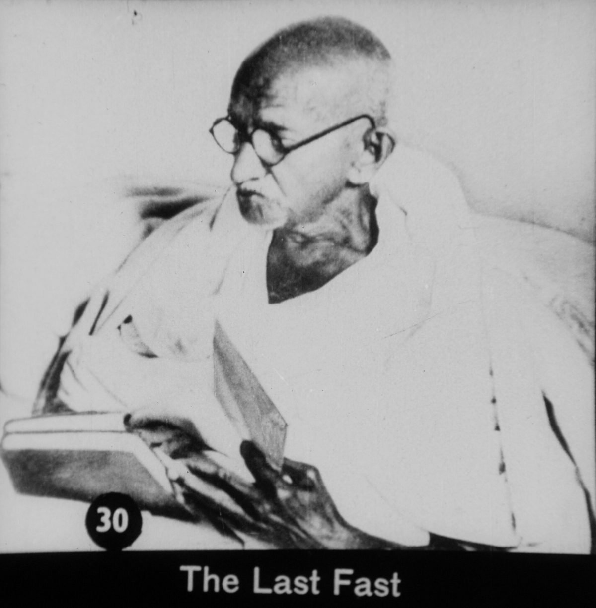 Black and white photograph of Gandhi lying on a cot under a white sheet during his fast, with people seated around him