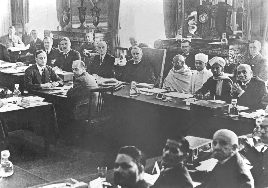 A long conference table in a panelled room with delegates in suits, Gandhi in white among them