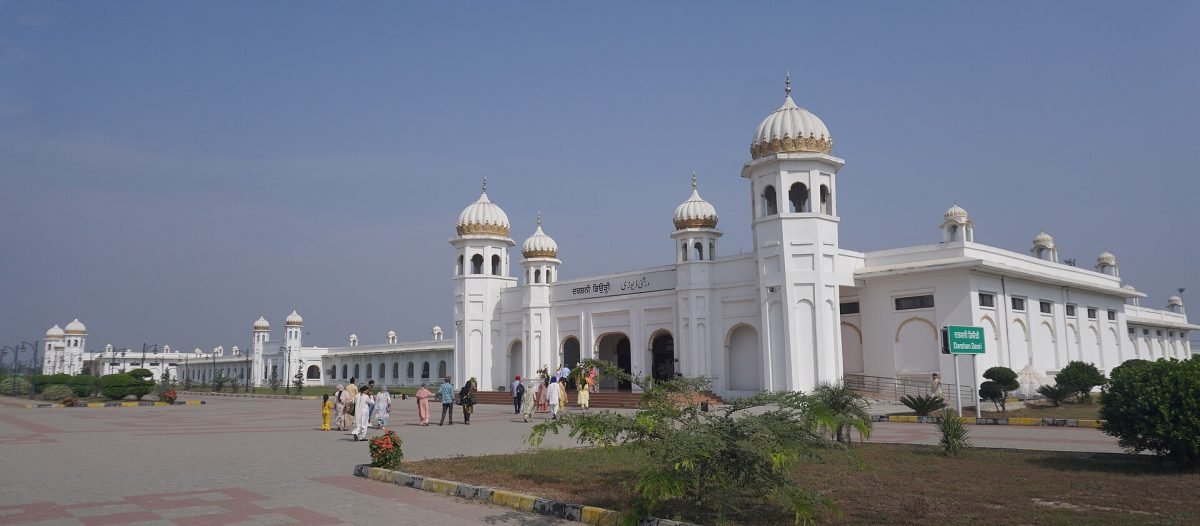 A long white gurdwara with domed kiosks and a central dome, with visitors walking across a wide paved courtyard