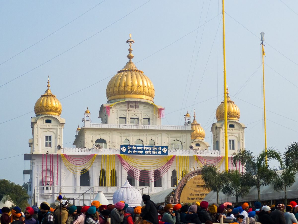 A white gurdwara with a large golden dome and smaller golden domes, decorated with saffron and pink cloth, with a large crowd in turbans and scarves in front