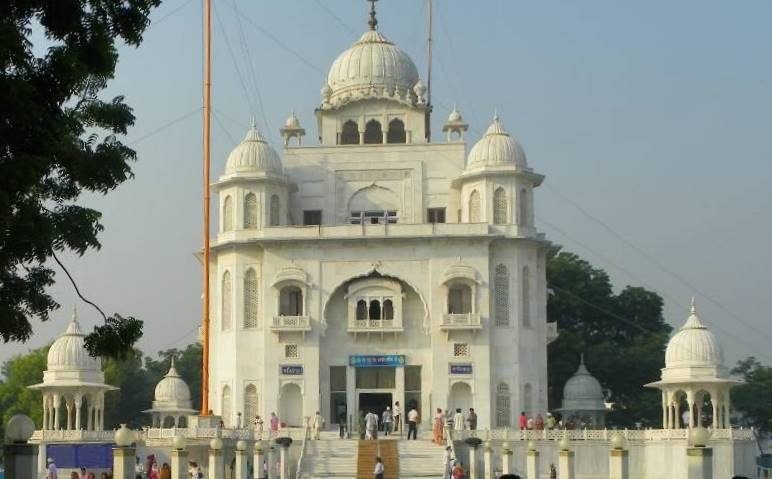 A large white gurdwara with domes behind a garden and lawns, under a clear sky