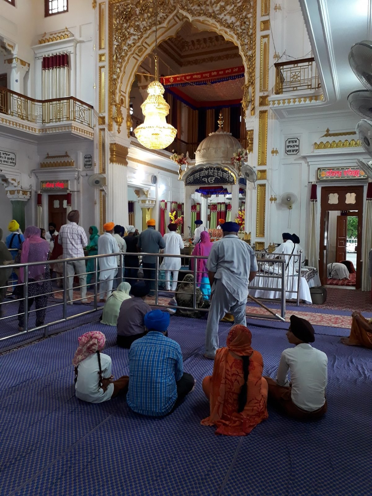 Worshippers sitting on a blue carpet inside a white and gold gurdwara hall, facing a canopied palki under a chandelier