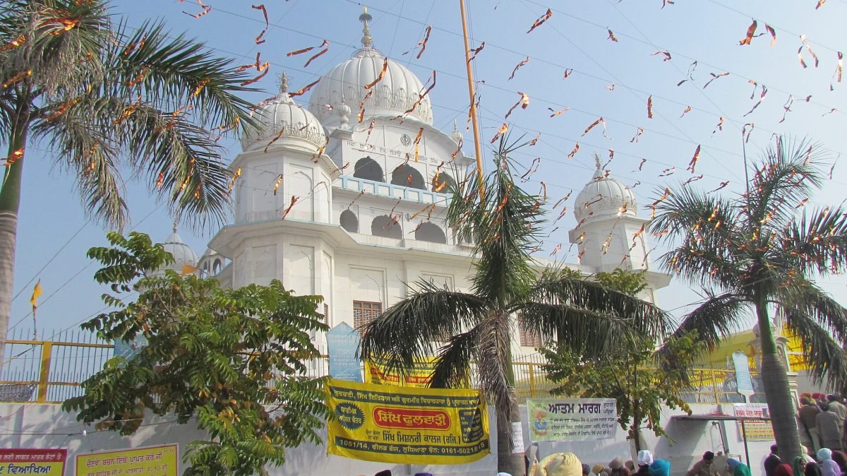 A white gurdwara with domes behind palm trees, with strings of small saffron flags overhead and pilgrims below