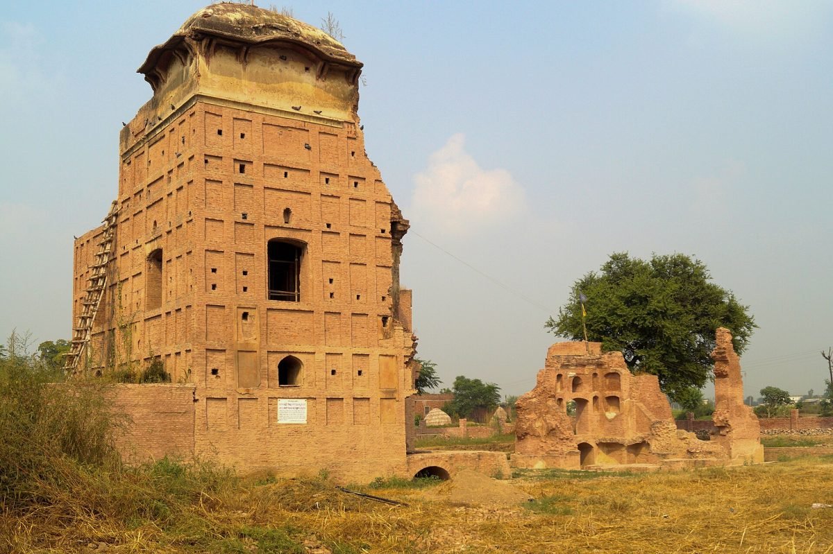A tall ruined brick building with empty window openings standing in dry grass, with crumbling walls beside it