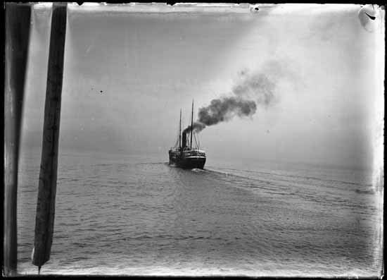 A black and white photograph of a steamship with a tall funnel moving across a harbour, with hills behind