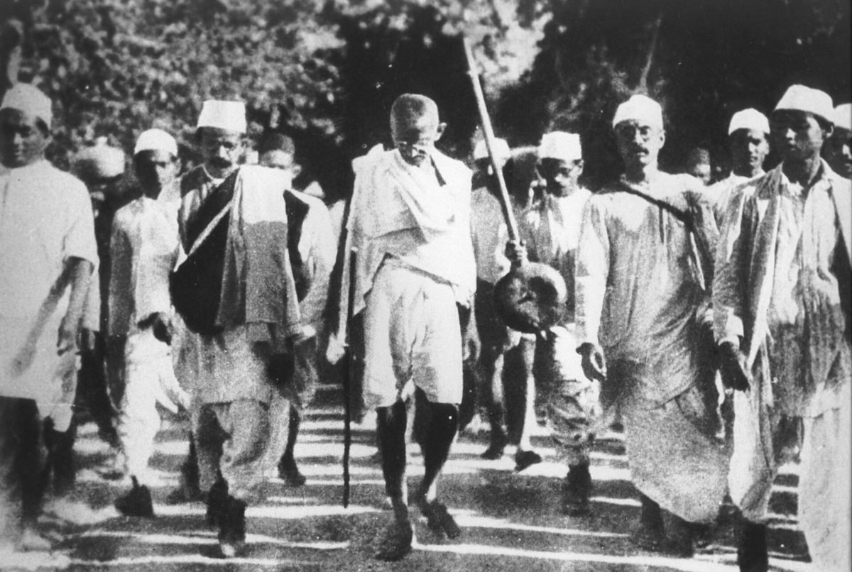 A black and white photograph of Gandhi, bare-chested with a white cloth and a long staff, walking at the head of a crowd of men in white