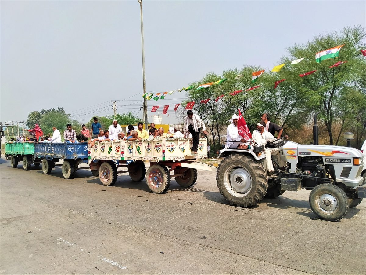 A white tractor pulls three trolleys full of farmers along a road strung with Indian tricolour flags and red flags