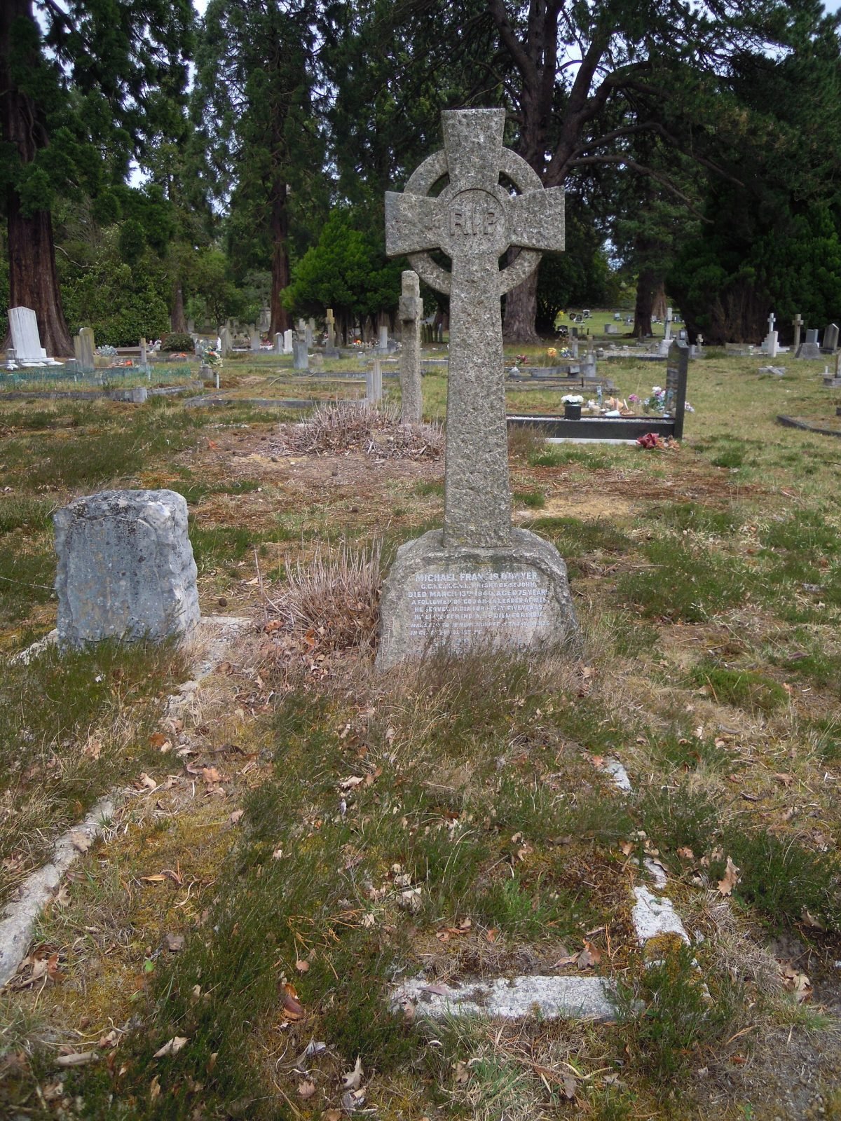 A weathered stone cross and grave marker in a grassy cemetery among trees