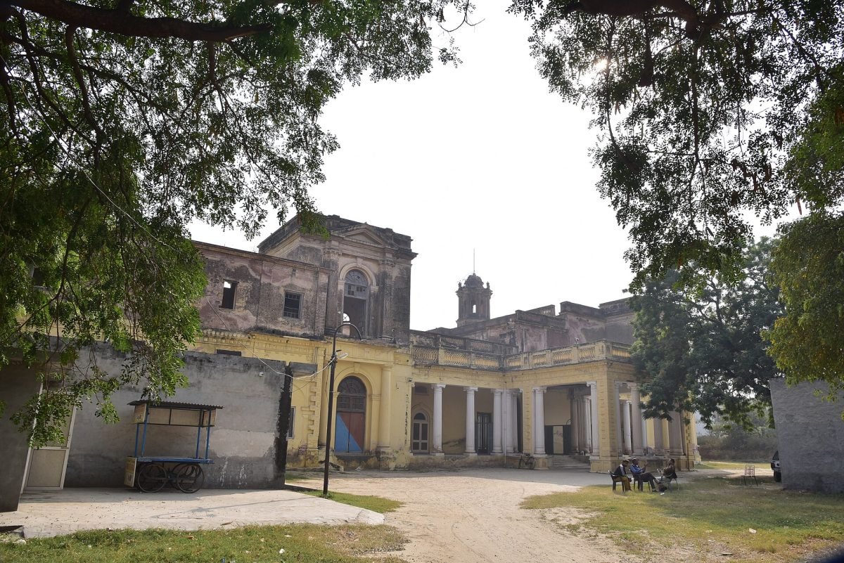 A weathered two-storey palace with columns and arched windows, seen through trees across a courtyard