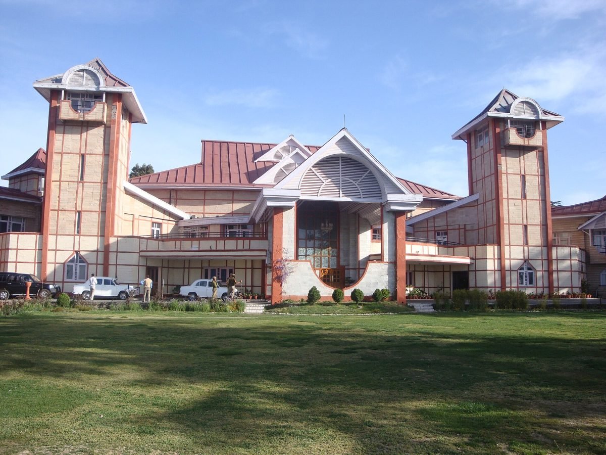 A large white colonial-style building with a sloping red roof among pine trees on a hillside
