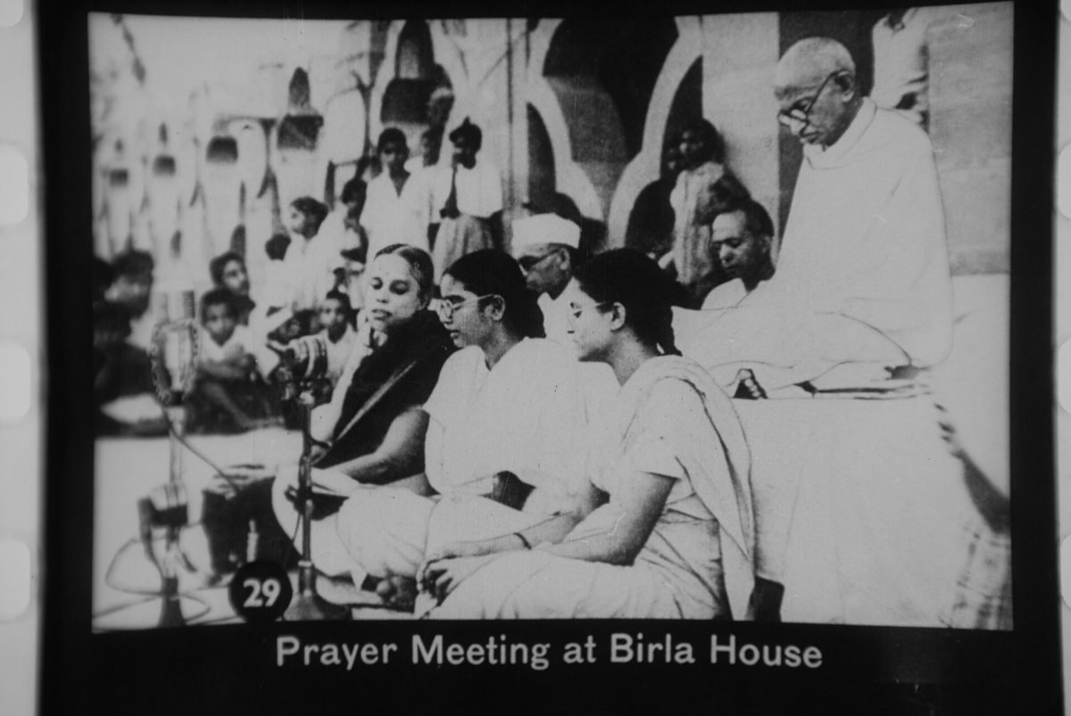 An old black and white photograph of a crowd seated on the ground in a garden at an outdoor prayer meeting, with Gandhi among them