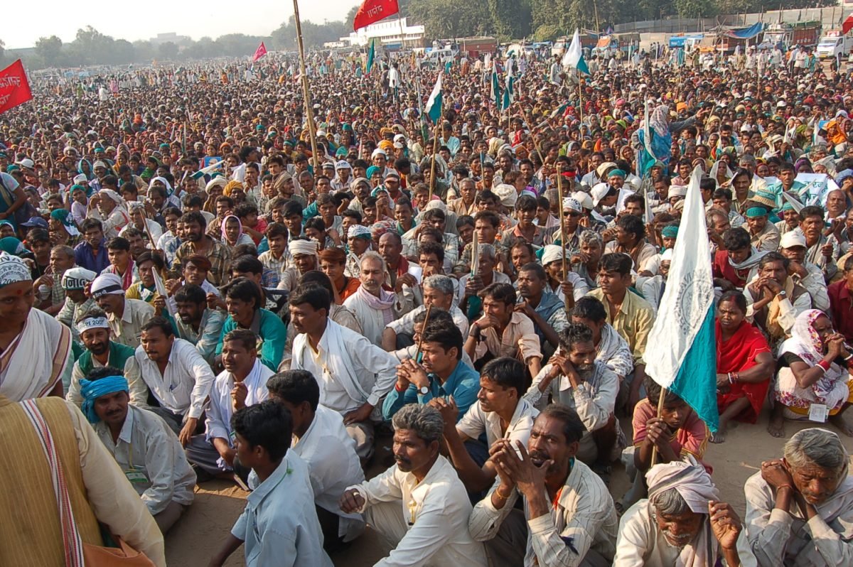A huge crowd seated on the open ground of Ramlila Maidan in Delhi, with banners and a stage