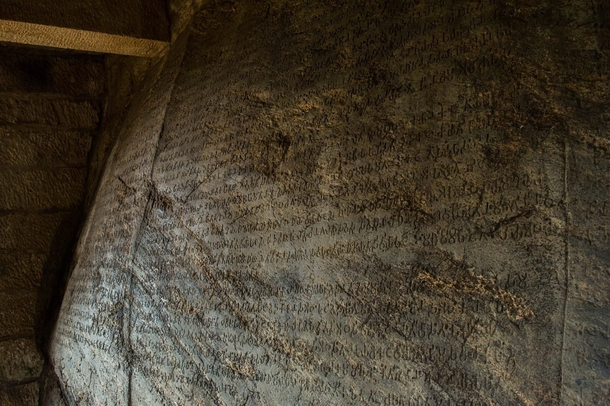 A large grey boulder on a hill with lines of ancient inscription cut into its face and the front of a carved elephant emerging from the top
