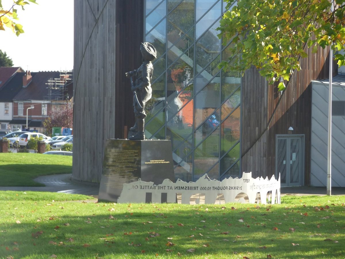 A bronze statue of a Sikh soldier on a stone plinth in a park, with a cut-out steel plate in front showing hills and lettering