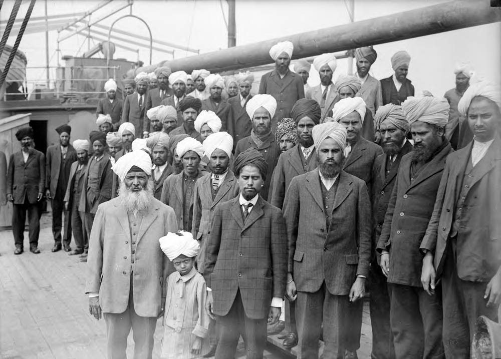 A black and white photograph of a crowd of turbaned men on the deck of a ship, with an older bearded man in a light suit in the left foreground