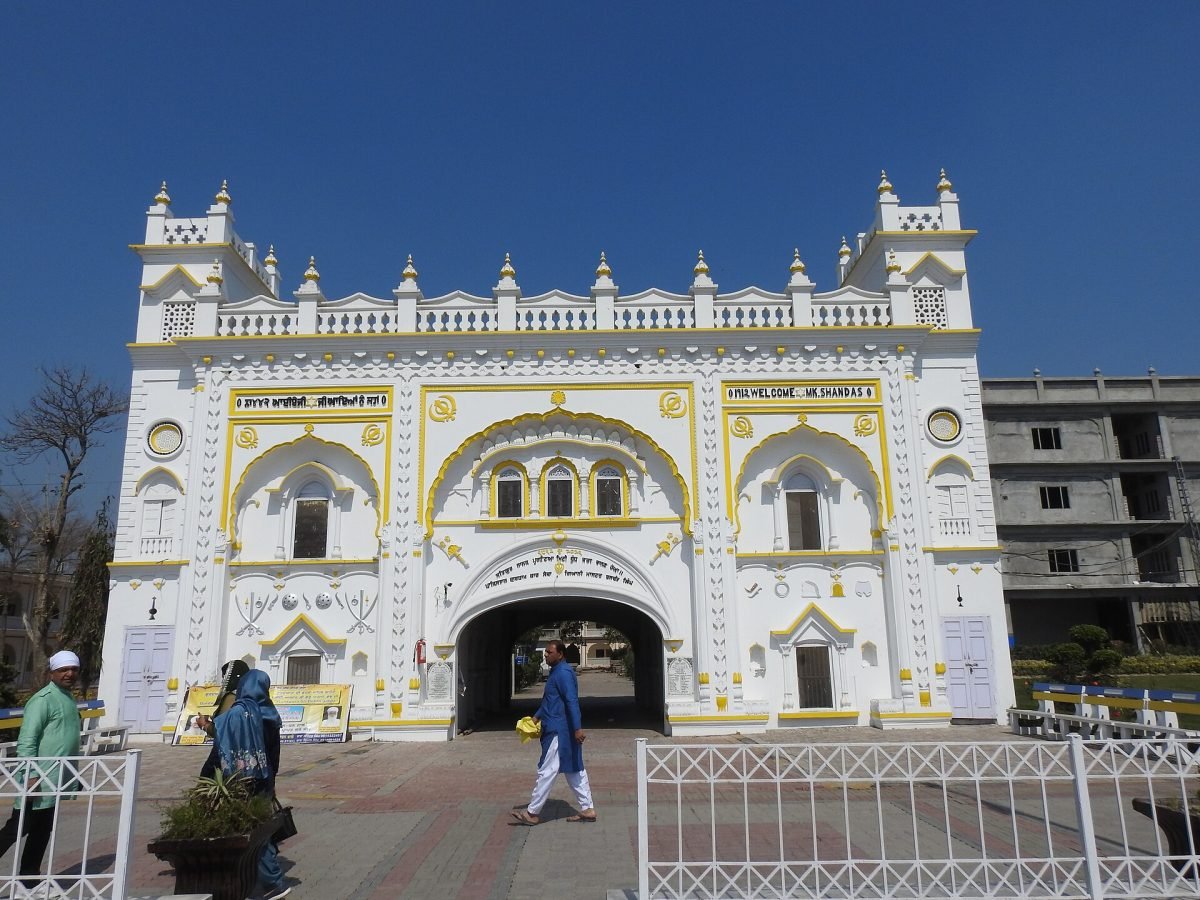 A white gateway with yellow trim and a tall arched entrance, with a few visitors walking in front of it under a blue sky