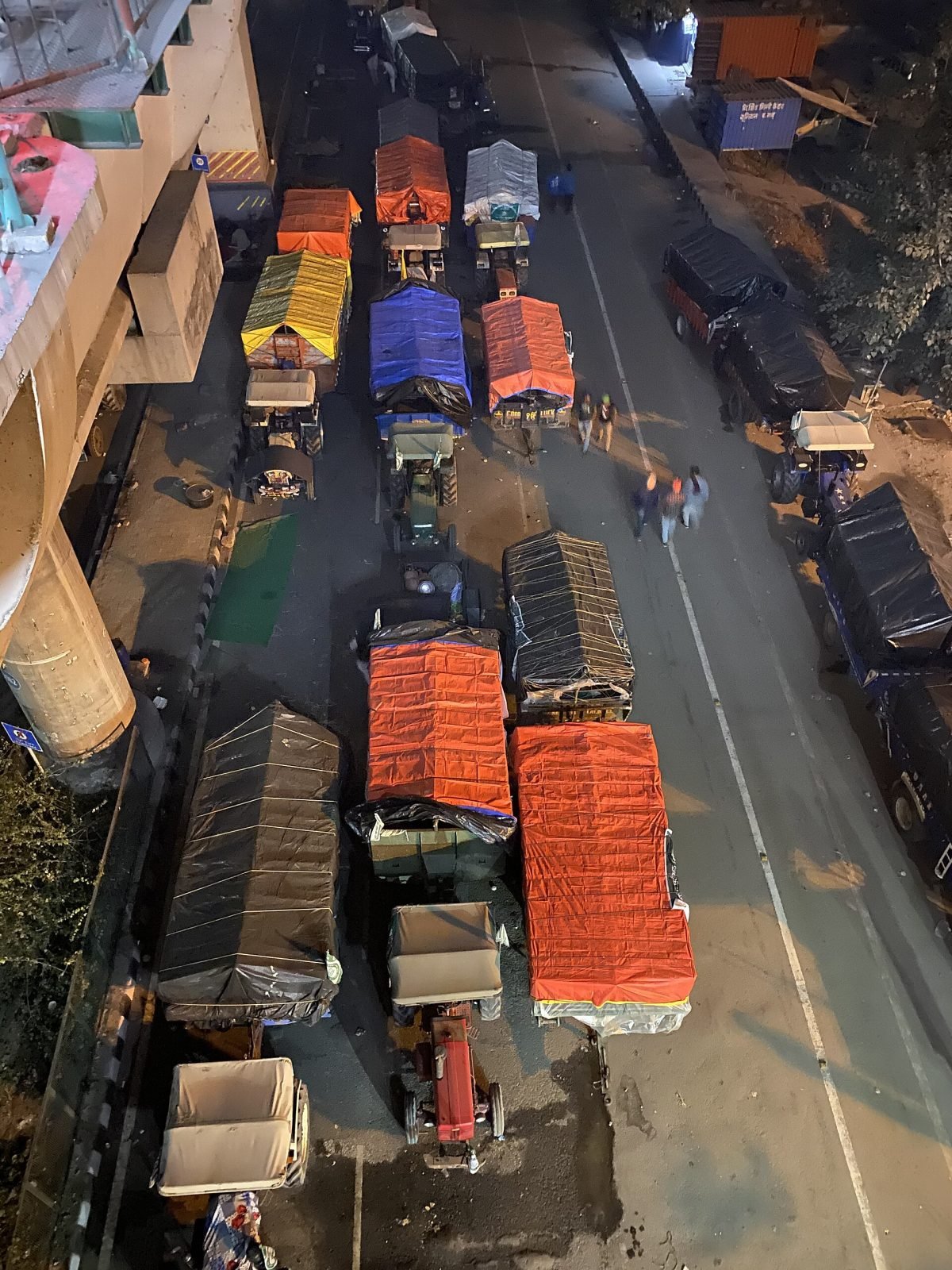 Rows of tractor trolleys covered in orange, blue and black tarpaulin parked on a road under a metro line at night