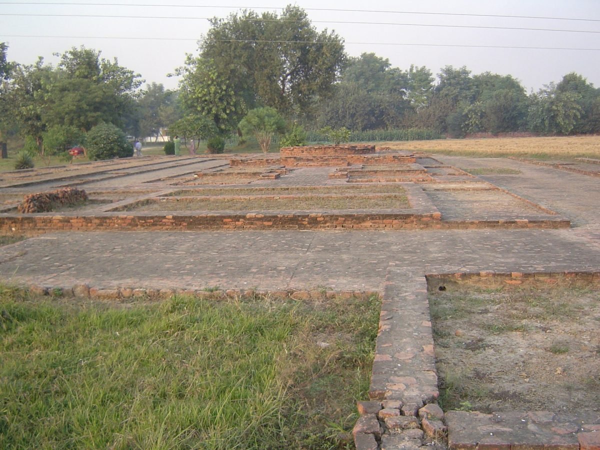 Low brick foundations of an excavated ancient site in a grassy field, with trees behind