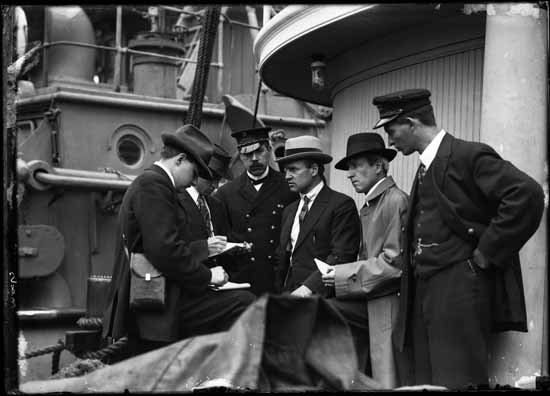 A black and white photograph of a group of men in suits and hats on a deck, talking with reporters