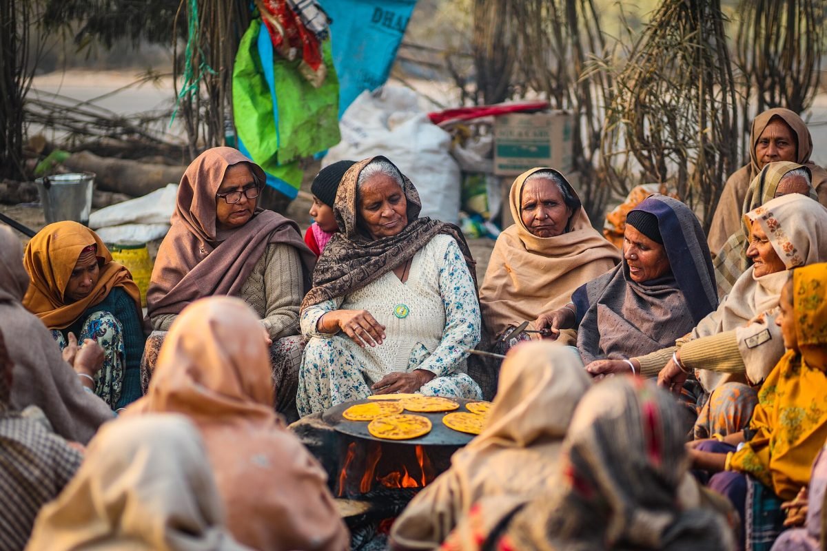 A group of older women in shawls sit around a large griddle over a fire, making rotis